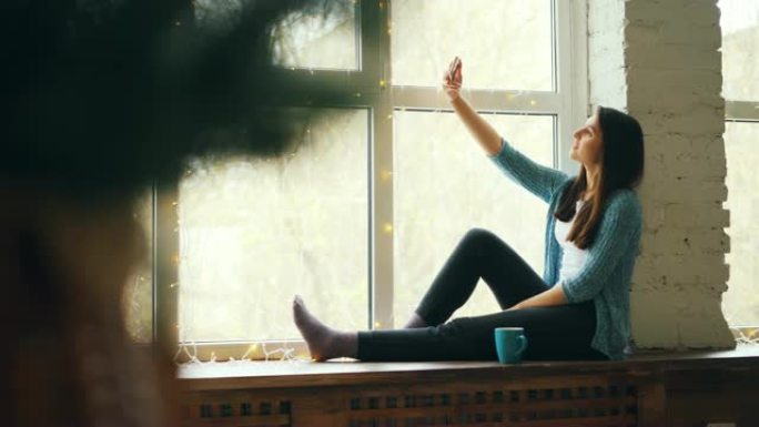 开朗的年轻女士正坐在窗台上进行在线视频通话，看着智能手机屏幕微笑着挥手交谈。通信和技术概念。