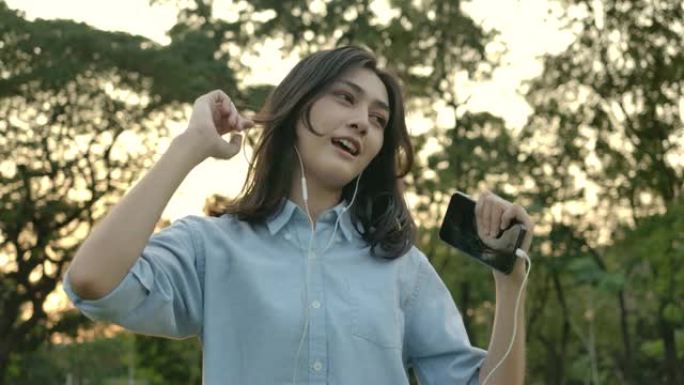 年轻迷人的亚洲女性在夏季公园里用电话和耳机听音乐，欣赏声音舞蹈。具有幸福情感概念的女人。慢动作。