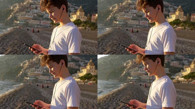 十几岁的男孩通过智能手机听音乐，在海滩上放松。穿着白色t恤的快乐男孩在意大利阿马尔菲波西塔诺享受日落