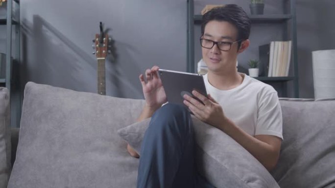 亚洲人坐在家里的客厅沙发上放松时，在数字平板电脑上阅读新闻。戴眼镜的成年男子在网上看报纸，在他的房子