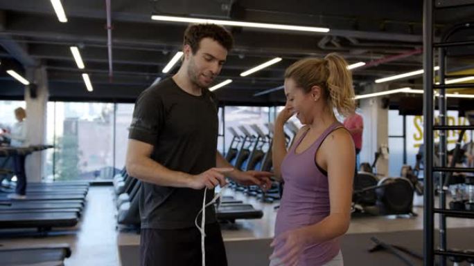 男性私人教练测量在健身房训练的年轻女子的手臂和腰部