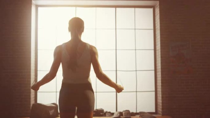 强壮的运动女性在阁楼风格的工业健身房用跳绳锻炼。她专注于激烈的交叉健身训练项目。面向大窗户。回击。