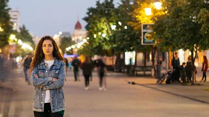 深夜，不幸的女孩独自站在美丽的步行街上，当人们四处呼啸时看着相机。社会与孤独概念。