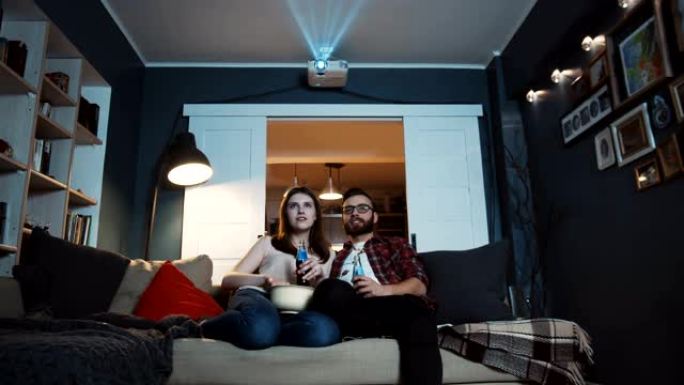 快乐的年轻高加索男人和女人坐在一起，用投影仪慢动作在家里看有趣的电影和零食。