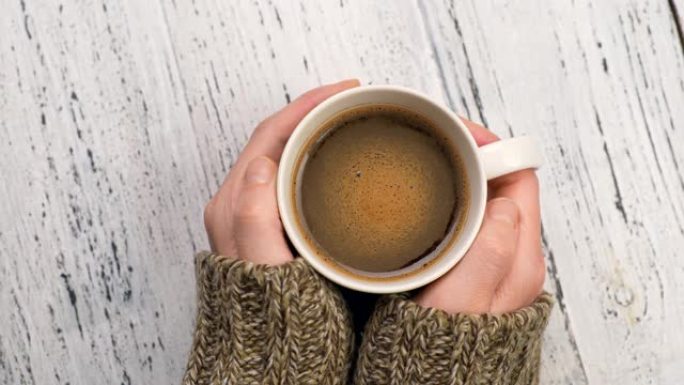 女人喝咖啡，温暖双手。穿着毛衣的女性手拿着一杯早间咖啡。破旧的白色乡村背景