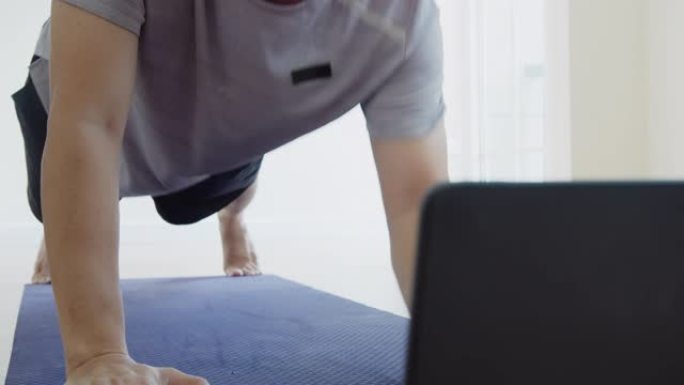亚洲男子在家里的客厅做肘板上下运动，通过笔记本电脑在线观看直播或视频教程。检疫期间的活动和社会距离新