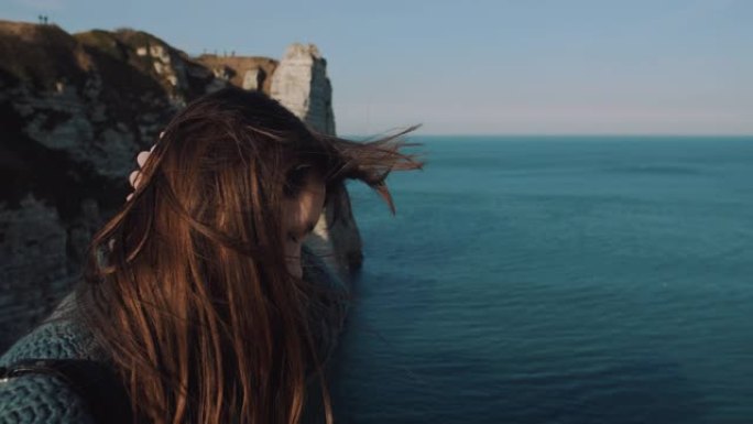 快乐的黑发旅游女人在埃特雷塔岩石海岸观看史诗般的景色，抚摸着在风中吹动的头发。