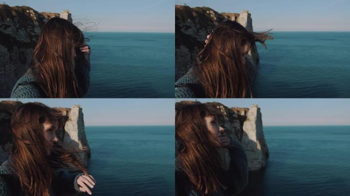 快乐的黑发旅游女人在埃特雷塔岩石海岸观看史诗般的景色，抚摸着在风中吹动的头发。