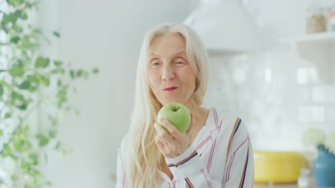 在家里明亮的厨房里吃苹果的高级女性养老金领取者的真实肖像。美丽的白发老女性对着镜头摆姿势，轻轻地微笑