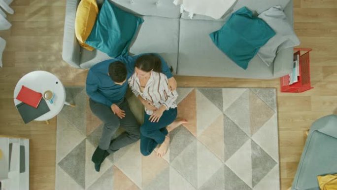 年轻夫妇坐在地板上聊天。他们进行对话。舒适的客厅，配有现代室内地毯、沙发、椅子、茶几、书架、植物和木