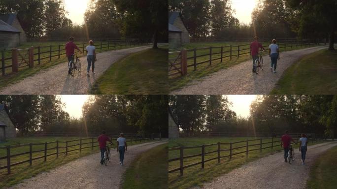 日落时分，浪漫情侣沿着乡间小路散步和推自行车的无人机后视镜头