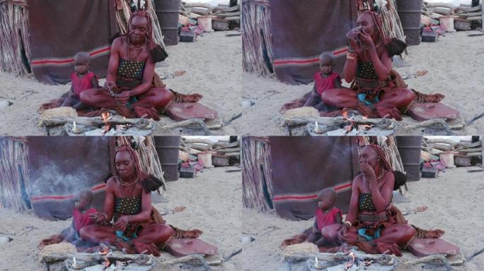 4K辛巴妇女的侧视图，穿着传统服装，带着年幼的孩子，在他们的小大院里的小屋外装满烟斗，吸烟，纳米比亚