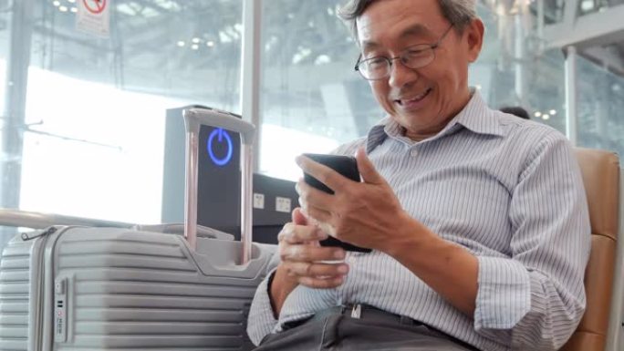 亚洲高级男子看着手机屏幕微笑。他坐在机场。旅行，人，旅程，全球，生活方式，假期，技术概念。在机场
