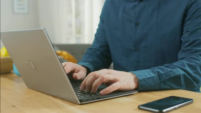 一个男人坐在家里的桌子旁，在他的现代银色笔记本电脑上工作的特写镜头。智能手机躺在他旁边的一张木桌上。