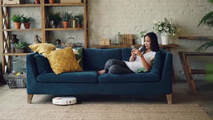 迷人的亚洲女孩正在享受现代技术，使用智能手机和机器人吸尘器坐在家里的沙发上。创新和室内设计概念。