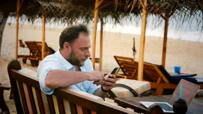 成功的男性自由职业者游客使用智能手机购物应用程序，同时坐在夏季海洋度假村沙滩椅上。