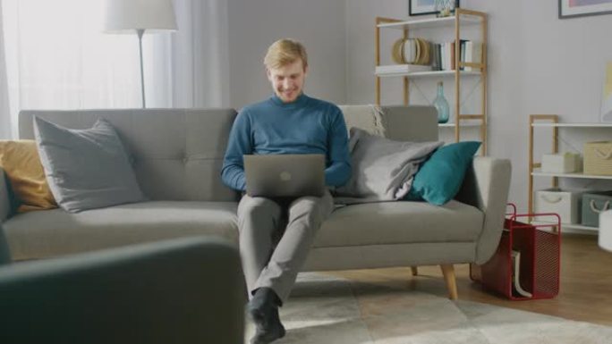 英俊的金发小伙子在笔记本电脑上工作的肖像，而坐在他舒适的客厅的沙发上。创意自由职业者在家放松，浏览互