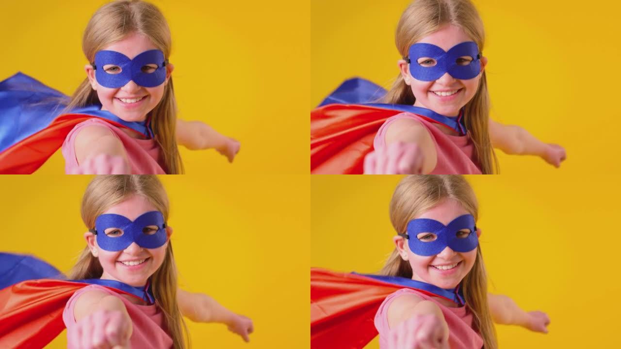 工作室拍摄的女孩打扮成漫画超级英雄戴着面具和斗篷假装在黄色背景下飞翔-慢动作拍摄
