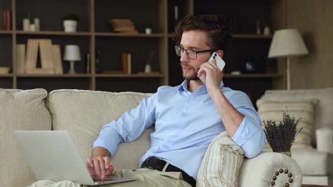 千禧一代的男人坐在沙发上使用笔记本电脑通过电话交谈