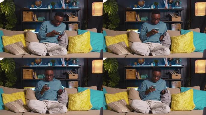 快乐有趣的黑人男学生在家里沙发上使用智能手机手机应用程序对音乐玩游戏点头。