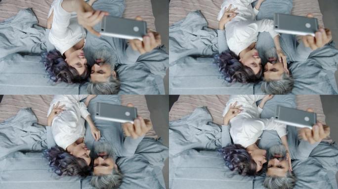 大胡子男人在床上用智能手机相机和漂亮的妻子自拍玩得开心