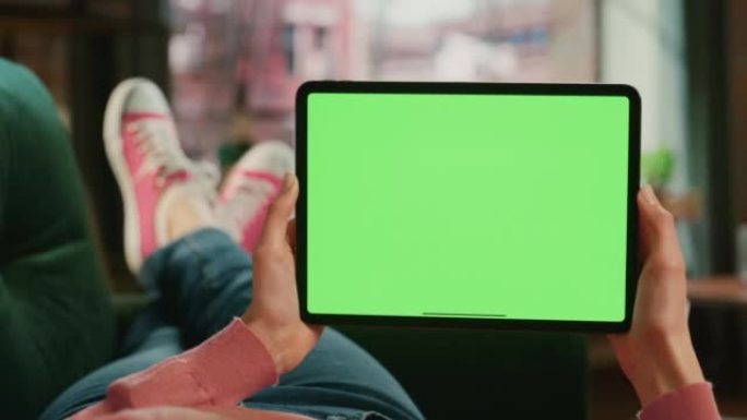 女性的手水平拿着一个绿屏显示的平板电脑。女性正在家里的沙发上放松，在移动设备上观看视频和阅读社交媒体