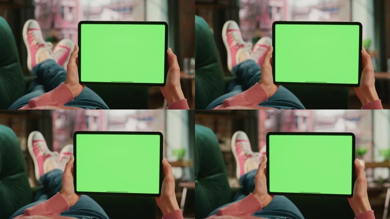 女性的手水平拿着一个绿屏显示的平板电脑。女性正在家里的沙发上放松，在移动设备上观看视频和阅读社交媒体