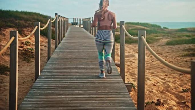 慢动作，日落时在海滩上的木制人行道上奔跑的女黑发运动员的跟踪镜头