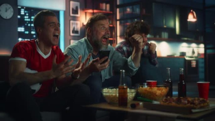 晚上在家:三个球迷坐在沙发上看电视比赛，使用智能手机应用程序在线打赌，当运动队获胜时庆祝胜利。吃零食