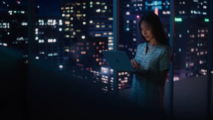 金融分析师使用笔记本电脑，站在窗户旁边，美丽的夜晚城市天际线与摩天大楼。自信的亚洲女商人在现代公司办
