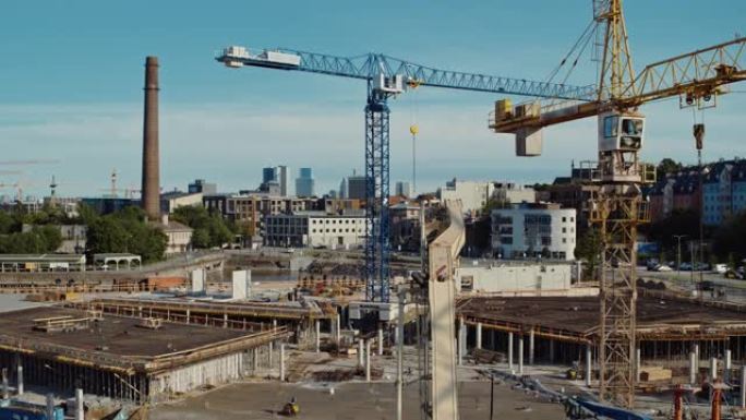 高空飞行在新的建筑开发场地上，用高塔起重机建造房地产。雇用重型机械和建筑工人。背景中的城市房屋。