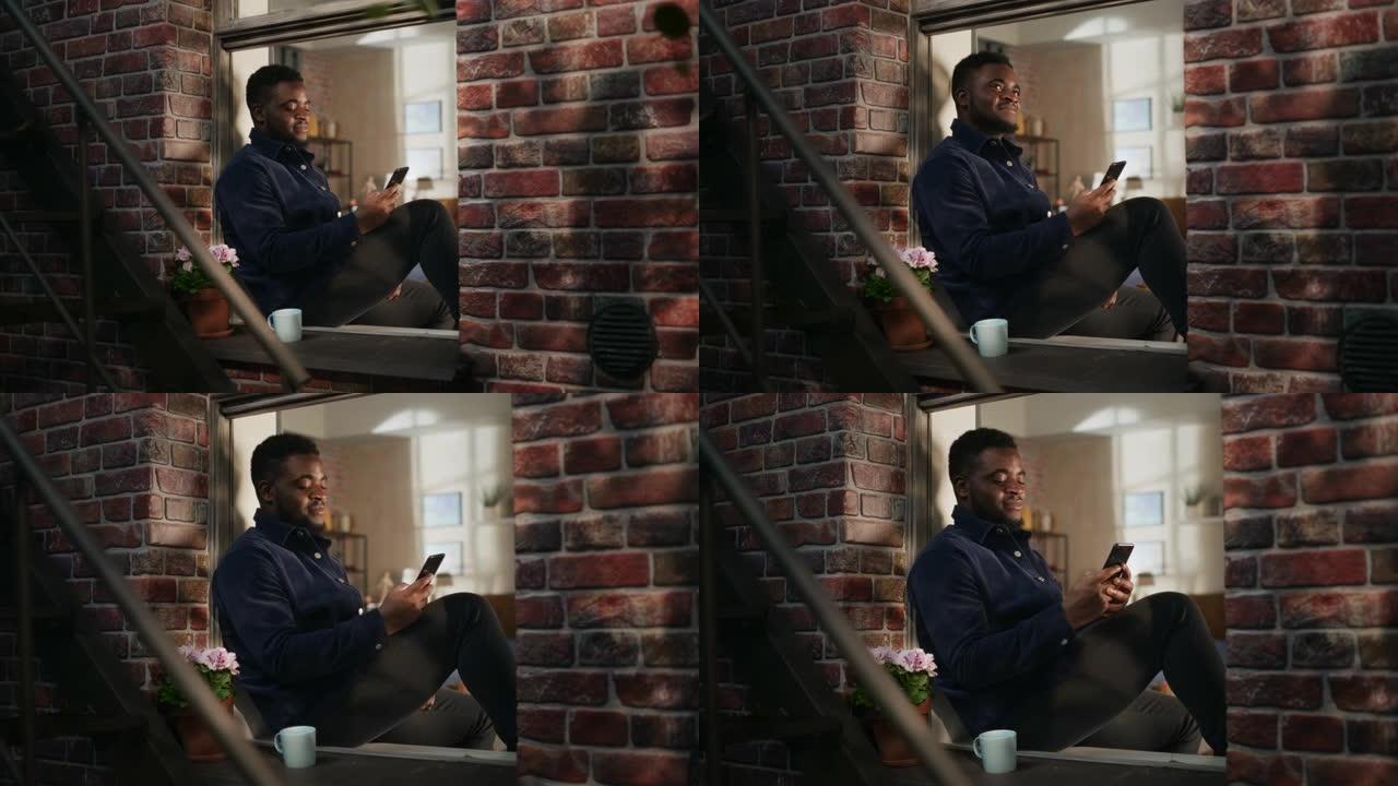 穿着舒适休闲服的黑人男性使用智能手机坐在窗台上。英俊的年轻人浏览互联网，一边微笑，一边看有趣的视频，