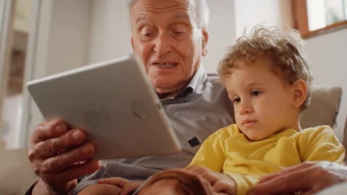 一个老人坐在沙发上，用平板电脑给他的小孙子读一本儿童读物的慢动作肖像。可爱的孩子仔细听他祖父讲的故事