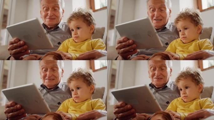 一个老人坐在沙发上，用平板电脑给他的小孙子读一本儿童读物的慢动作肖像。可爱的孩子仔细听他祖父讲的故事