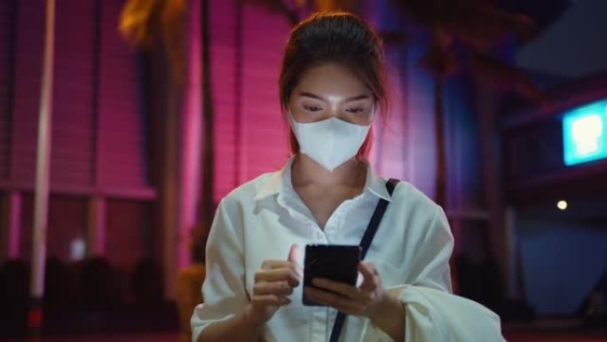 年轻的亚洲女商人穿着时尚的办公室衣服，戴着医用口罩，使用智能手机打字短信，晚上站在城市现代城户外。