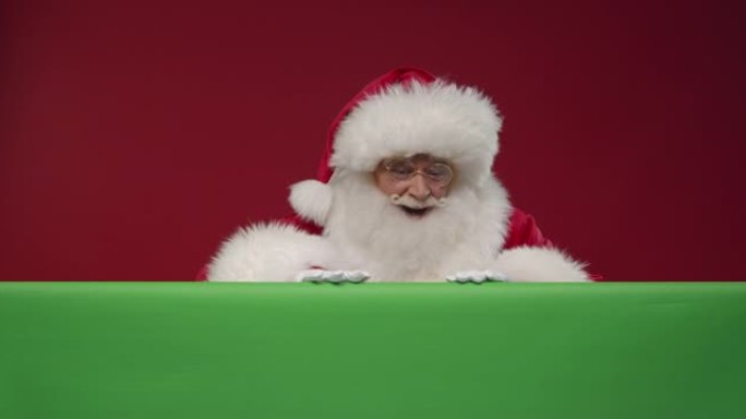 圣诞老人从他前面的绿色屏幕后面出现在红色背景的框架中，并显示拇指向上的标志