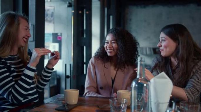 电影拍摄的三个美丽的年轻快乐的多民族女性朋友微笑着，在时尚的咖啡桌旁随意交谈。