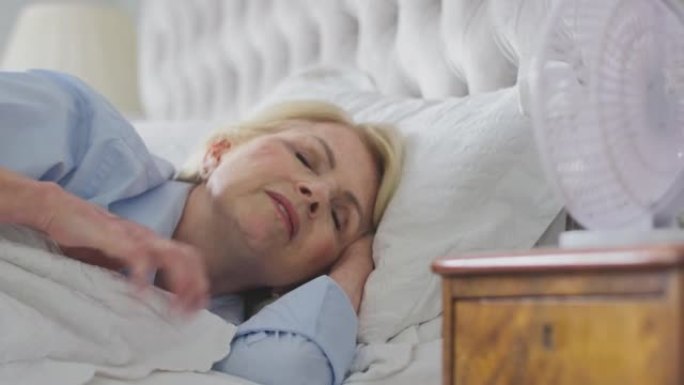 患有失眠和热潮红的更年期成熟女性在家床上用电风扇慢动作冷却自己