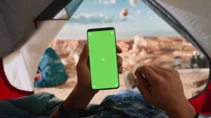 一个游客垂直拿着带有绿屏占位符的智能手机，用手指在显示屏上滑动的POV视图。旅行者在落基山顶上的帐篷