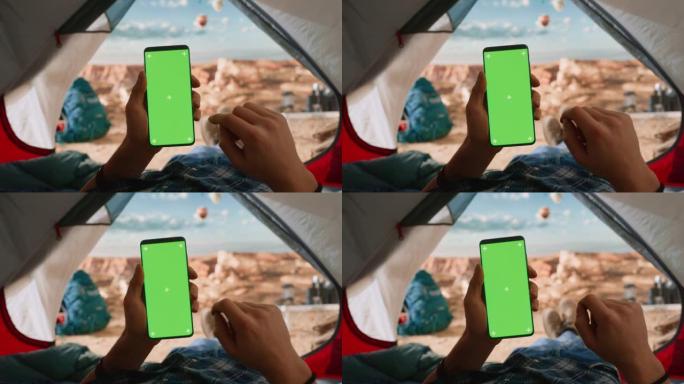 一个游客垂直拿着带有绿屏占位符的智能手机，用手指在显示屏上滑动的POV视图。旅行者在落基山顶上的帐篷