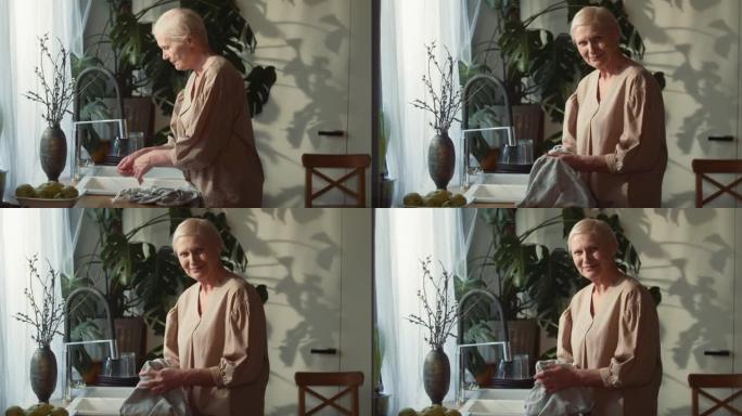 美丽的60-65岁高年级男子肖像微笑的白人妇女洗净，在厨房水槽用毛巾擦干手，看着相机。