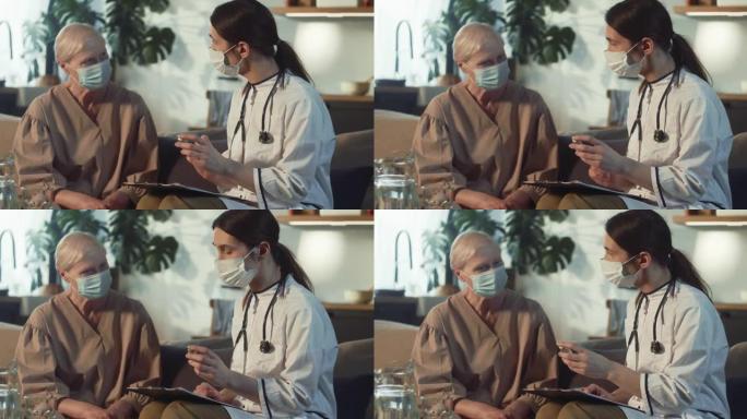 穿着实验室外套的年轻女医生在家访期间向戴着医用口罩的老年妇女展示带有疫苗的药瓶。