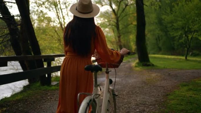 中等，跟踪镜头，一个无法辨认的黑发穿着橙色连衣裙，在河边的森林里骑自行车，慢动作