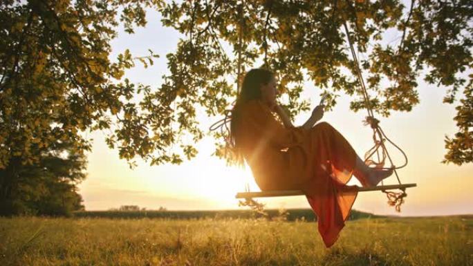 一位穿着红色连衣裙的年轻成年女性的静态镜头摇摆着看着手机，坐在悬挂在大树枝上的绳索秋千上，慢动作
