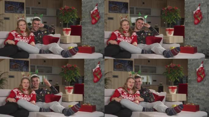 笑着的年轻夫妇坐在舒适的沙发上，在电视上看圣诞喜剧