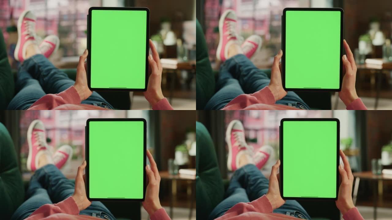 女性的手垂直拿着一个绿屏显示的平板电脑。女性正在家里的沙发上放松，在移动设备上观看视频和阅读社交媒体