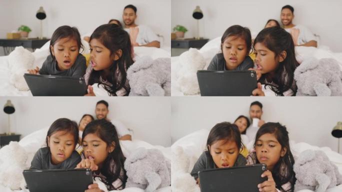 小女孩与父母坐在床上，在数字平板电脑上学习和浏览。父母看着女儿在线播放和播放视频。年轻的家庭一起放松