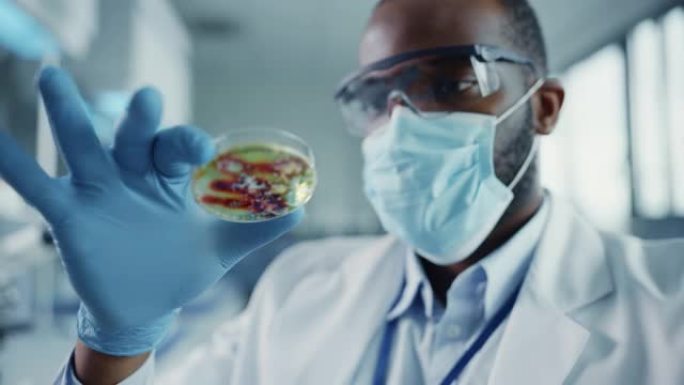 非裔美国男性科学家戴着口罩和眼镜看着带有转基因样品化学物质的培养皿。在具有技术设备的现代实验室中工作