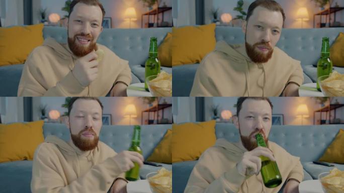 迷人的大胡子男人在发光的房子里吃薯条喝啤酒，嘲笑电视上的有趣电影的肖像