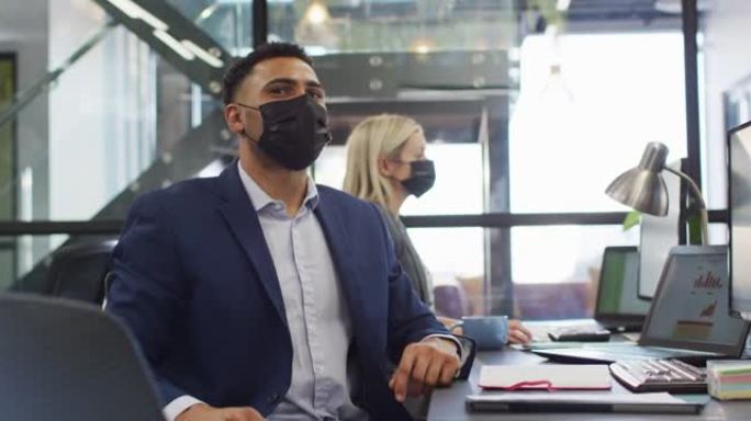 混血商人戴着口罩并与同事在后台工作时使用计算机